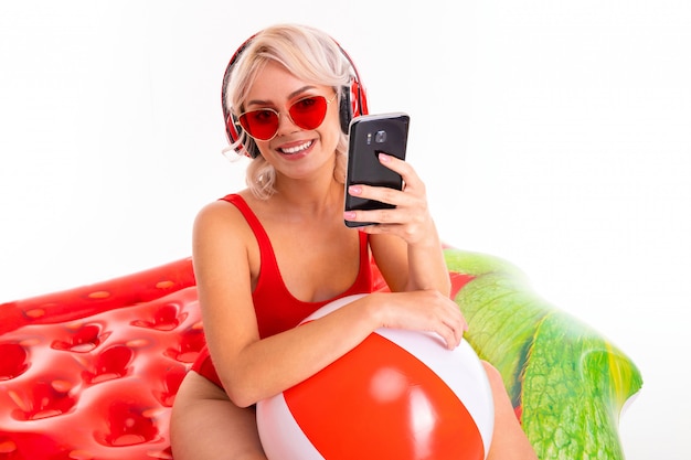 jeune fille blonde dans un maillot de bain rouge et des lunettes de soleil assis sur un matelas de natation et écouter de la musique sur un casque et tenant un téléphone dans ses mains