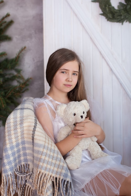 La jeune fille blonde dans une robe blanche est assise sur une chaise dans une étreinte avec un ours en peluche et rêve. Maison confortable. Chambre éco-style en harmonie avec la nature. Ambiance de Noël, confort.