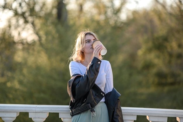 Une jeune fille blonde dans une veste en cuir boit du café en été