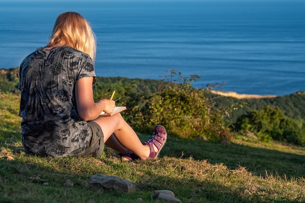 Une jeune fille blonde écrit ses pensées dans un journal personnel sur la montagne avec vue sur la mer, profitez de votre temps et de vos moments de voyage et de détente