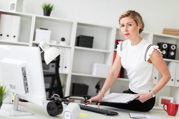 Une jeune fille blonde est assise sur un bureau dans le bureau. Elle tient des documents et un crayon à la main et imprime sur le clavier.