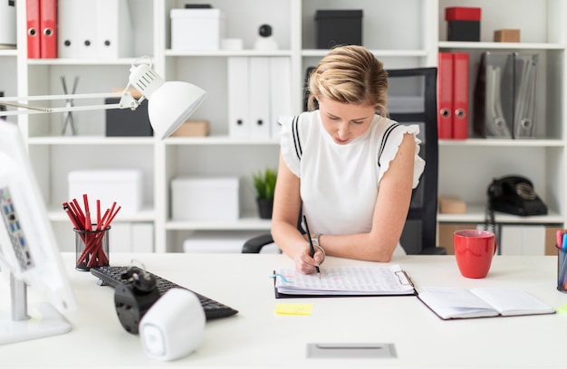 Une jeune fille blonde est assise à un bureau d'ordinateur dans le bureau et écrit un crayon dans le document.