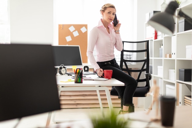 Une jeune fille blonde est assise sur une table dans le bureau, tient une tasse rouge dans sa main et son téléphone.