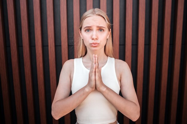 Jeune fille blonde est debout contre le mur et regarde. Elle garde ses mains en prière. Jeune femme est bouleversée et garde ses lèvres ensemble.