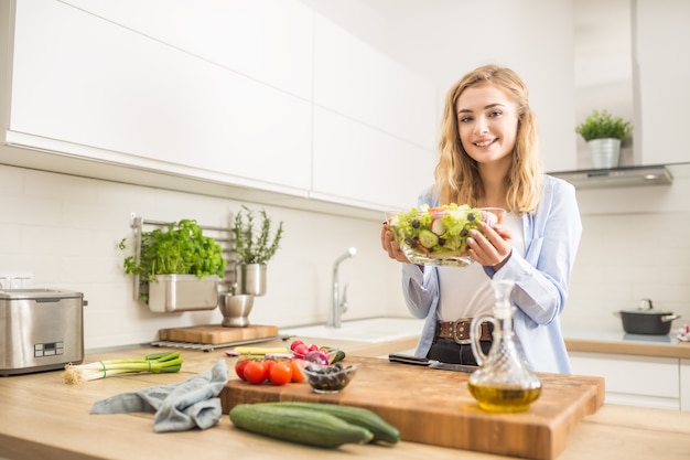 Jeune fille blonde heureuse préparant une salade saine dans la cuisine à domicile.