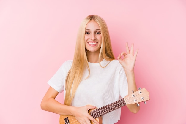 Jeune fille blonde jouant du ukelele joyeux et confiant montrant le geste ok.