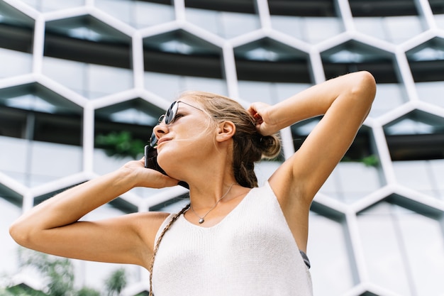 Jeune fille blonde avec des lunettes de soleil debout devant un immeuble moderne en parlant avec le mobile