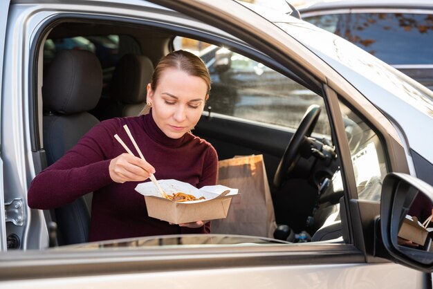 Photo une jeune fille blonde mangeant de délicieux nouilles chinoises dans la boîte avec des baguettes assises dans la voiture.