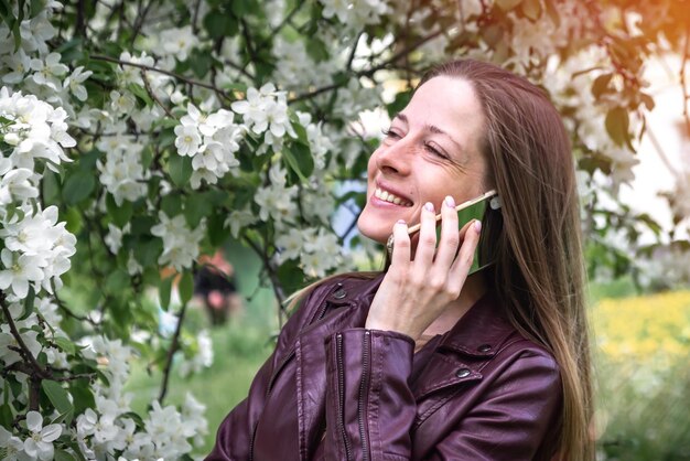 Une jeune fille blonde marchant dans le parc parle sur un smartphone et rit la saison du printemps est la floraison du pommier les gens utilisent la technologie