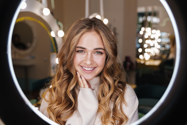 une jeune fille blonde mignonne heureuse à l'intérieur dans un salon de beauté regardant à l'avant à travers une lampe à lumière annulaire.