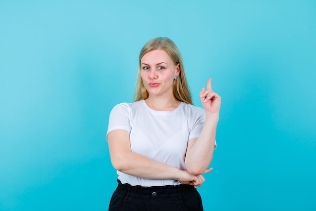 Jeune fille blonde pointe vers le haut avec l'index sur fond bleu