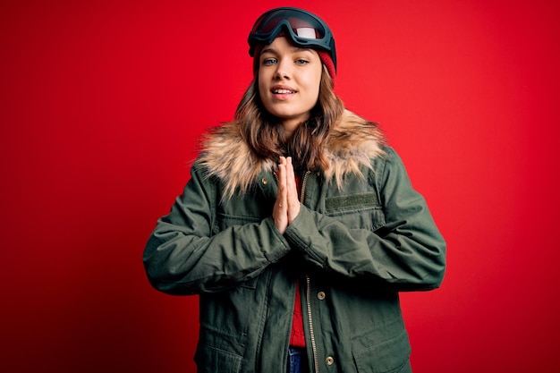 Photo jeune fille blonde portant des lunettes de ski et un manteau d'hiver pour le temps de ski sur fond rouge priant avec les mains ensemble pour demander pardon souriant confiant