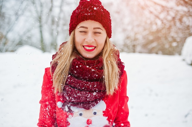 Jeune fille blonde en pull rouge écharpe, bonnet et santas posant au parc le jour de l'hiver.