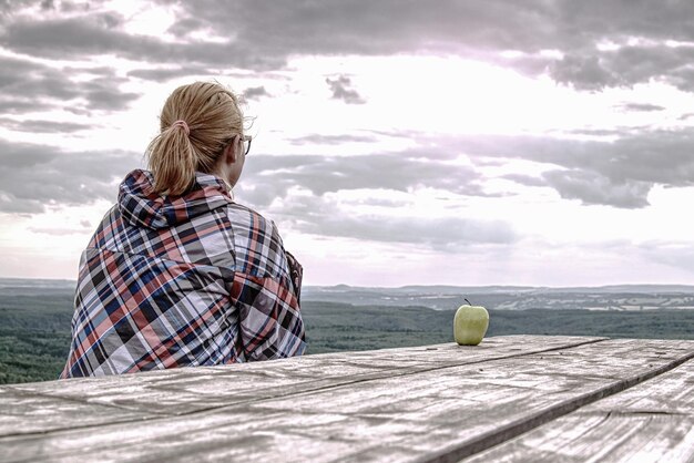 Photo une jeune fille blonde se repose à une table en bois et prend une pause de randonnée. une pomme verte sur la table.