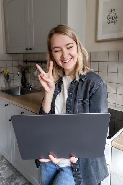 Jeune fille blonde se tient avec un ordinateur portable dans la cuisine et travaille à domicile