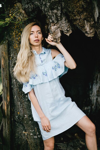 Jeune fille blonde séduisante en robe bleue posant avec vieil arbre