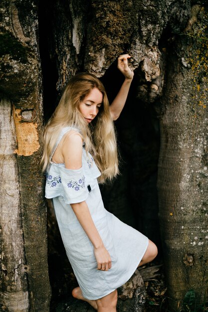 Jeune fille blonde séduisante en robe romantique bleue posant avec vieil arbre