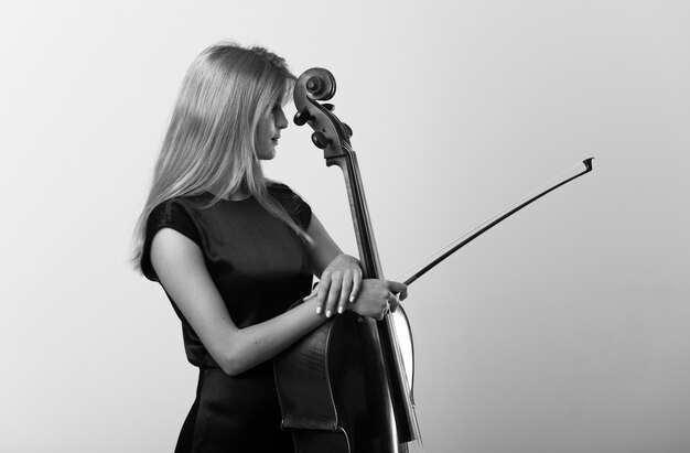 Jeune fille blonde avec son violoncelle posant sur un mur blanc