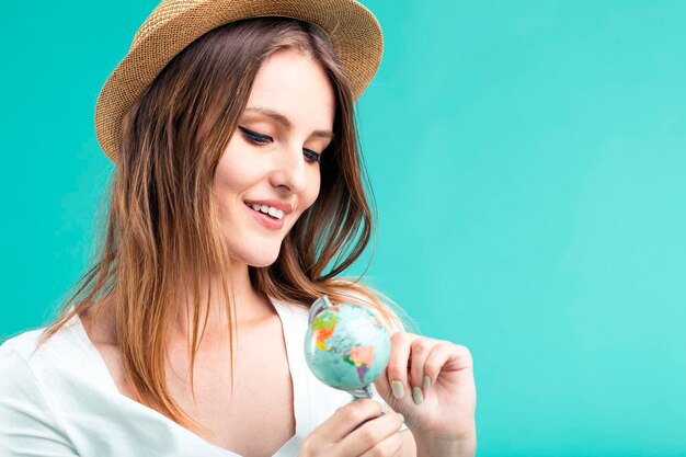 Jeune fille blonde souriante porte un t-shirt blanc et un chapeau tenant un petit globe sur fond bleu concept de destination de vacances d'été