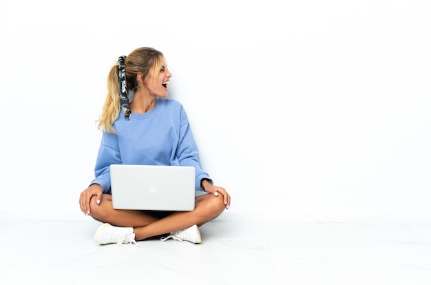 Jeune fille blonde uruguayenne avec l'ordinateur portable sur blanc en riant en position latérale