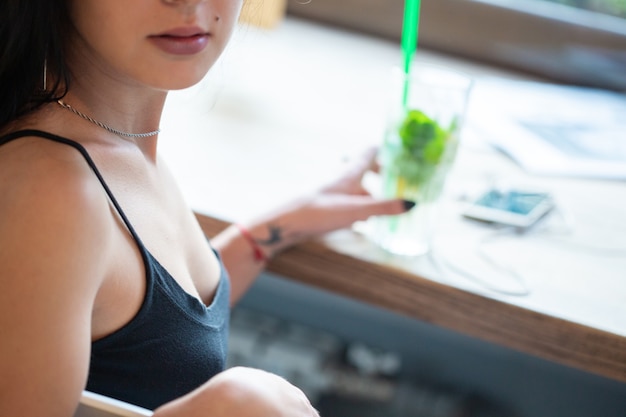 Une jeune fille boit un mojito avec une paille alors qu'elle est assise dans un café près de la fenêtre. Tiré du côté droit.