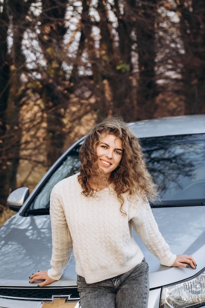 Photo une jeune fille bouclée dans un pull blanc sur le fond de la nature d'automne et de sa propre voiture