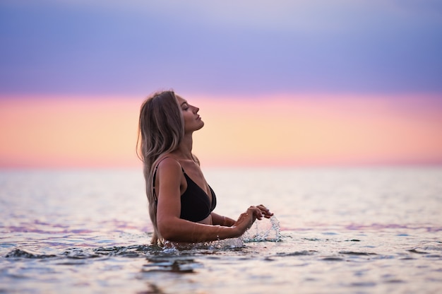 Une jeune fille bronzée mince avec des cheveux blonds luxuriants dans un maillot de bain noir, éclabousse sur les côtés alors qu'elle était assise dans un estuaire peu profond sur fond d'un coucher de soleil chaud et lumineux