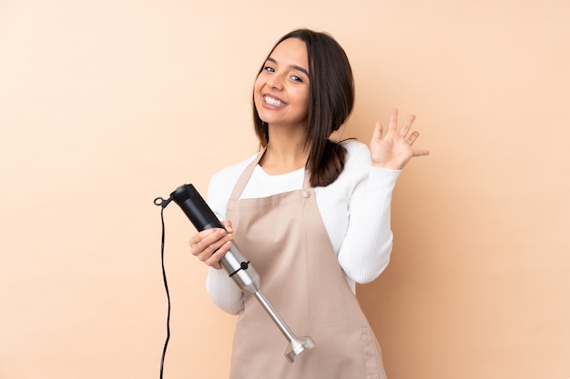 Jeune fille brune à l'aide d'un mélangeur à main sur un mur isolé saluant avec la main avec une expression heureuse