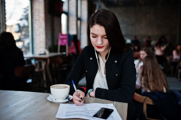 Jeune fille brune assise sur un café avec une tasse de cappuccino en écoutant de la musique au casque et en écrivant des documents.