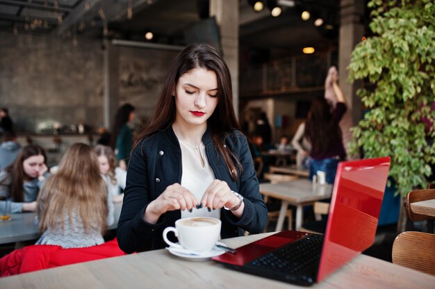 Jeune fille brune assise sur un café avec une tasse de cappuccino, travaillant avec un ordinateur portable rouge.