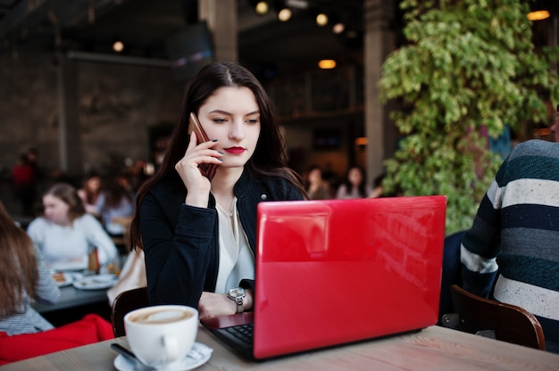 Jeune fille brune assise sur un café, travaillant avec un ordinateur portable rouge et parlant au téléphone mobile.