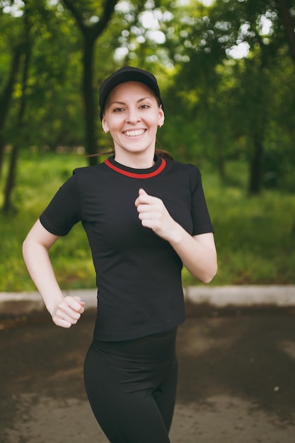 Jeune Fille Brune Athlétique Souriante En Uniforme Noir Et Entraînement De Casquette Faisant Des Exercices De Sport, Courant Et Regardant La Caméra Sur Le Chemin Dans Le Parc De La Ville à L'extérieur