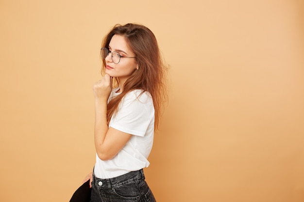 Jeune fille brune aux cheveux longs vêtue d'un t-shirt blanc et d'un jean noir pose avec un chapeau noir à la main sur le fond beige en studio.