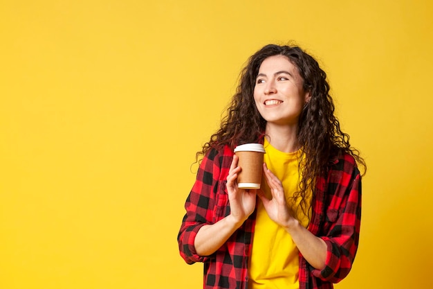 Jeune fille brune bouclée dans une chemise à carreaux tient du café sur un fond jaune