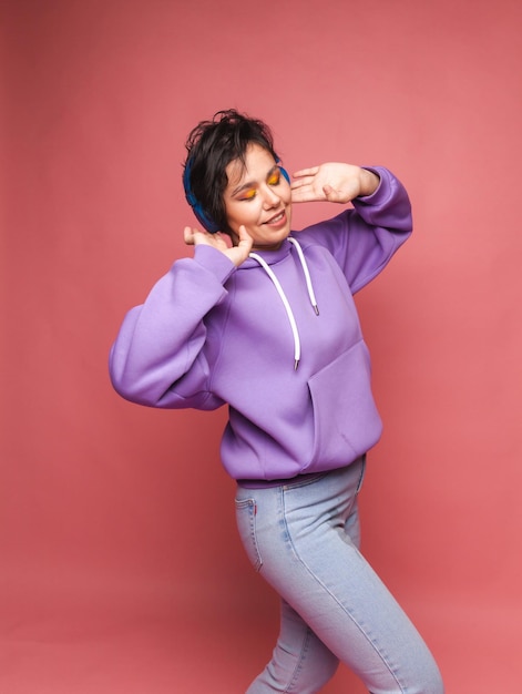 Jeune fille brune dans un pull lilas et un casque pose isolément sur un fond rose dans le studio Le concept de mode de vie des gensÉcouter de la musique avec un casque portrait de studio de danse