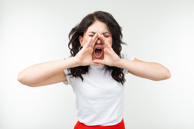 Jeune fille brune dans un T-shirt blanc criant de nouvelles bruyamment tenant ses mains près de sa bouche sur un fond blanc