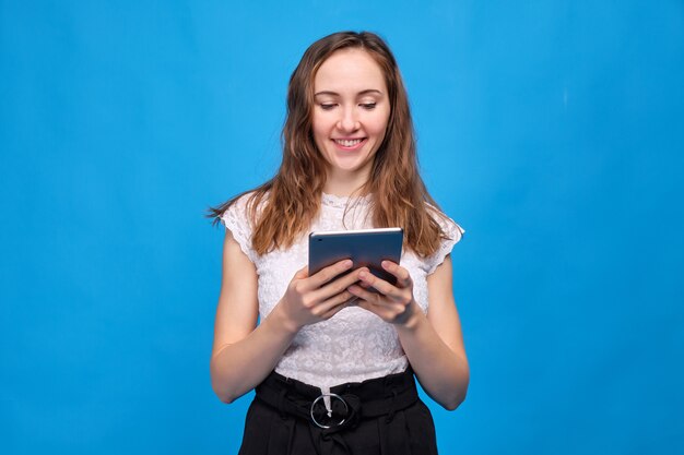 Jeune fille brune dans des vêtements décontractés sur fond bleu et regarder des films drôles, des émissions de télévision, des spectacles à travers une tablette