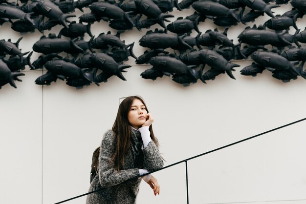 Une jeune fille brune élégante en manteau gris se tient dans les escaliers sur fond de mur inhabituel