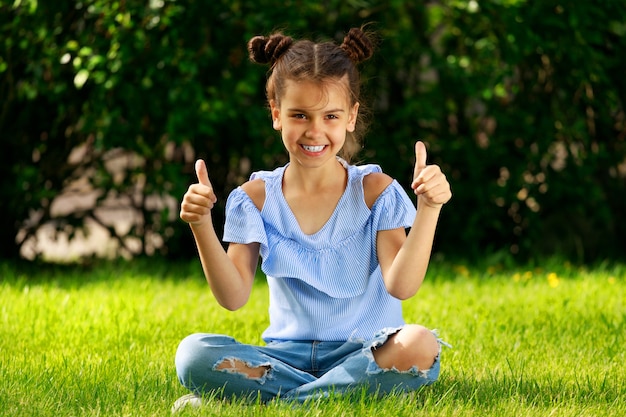 Jeune Fille Brune Est Assise Sur L'herbe Dans Le Parc Et Montre Ses Pouces. Photo De Haute Qualité