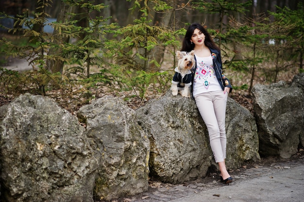 Jeune fille brune gitane avec chien yorkshire terrier posé contre des pierres sur le parc. Modèle porter sur une veste en cuir avec ornement, un pantalon et des chaussures à talons hauts.