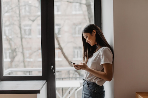 Jeune fille brune en jeans et haut blanc avec sourire regarde dans le téléphone s'appuyant sur la fenêtre