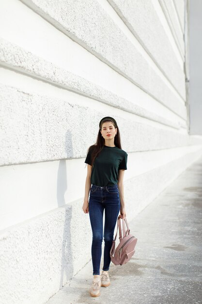 Jeune fille brune en jeans un T-shirt vert une bande de cheveux et un sac à dos rose se dresse contre un mur blanc