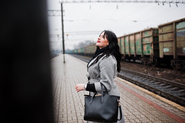 Jeune fille brune en manteau gris posée dans une gare