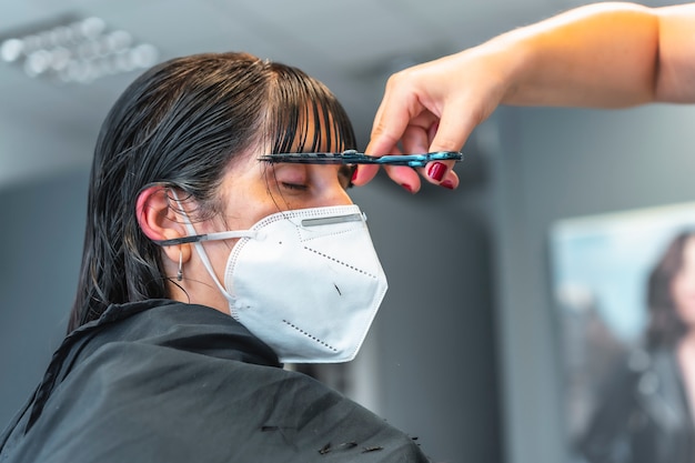 Jeune fille brune avec masque facial dans un salon de coiffure lui coupant la frange. Réouverture avec des mesures de sécurité pour les coiffeurs dans la pandémie de Covid-19. Nouvelle normale, coronavirus, distance sociale