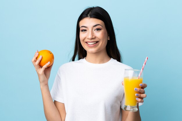 Jeune fille brune sur mur bleu isolé tenant une orange et un jus d'orange