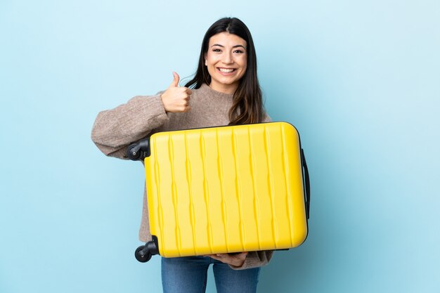 Jeune fille brune sur mur bleu isolé en vacances avec valise de voyage et avec le pouce vers le haut