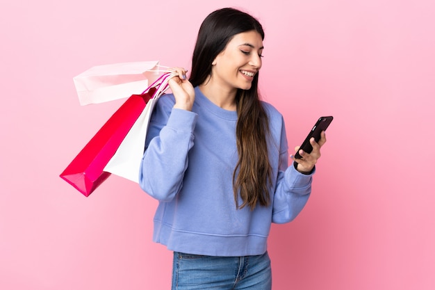 Jeune fille brune sur un mur rose tenant des sacs à provisions et écrivant un message avec son téléphone portable à un ami