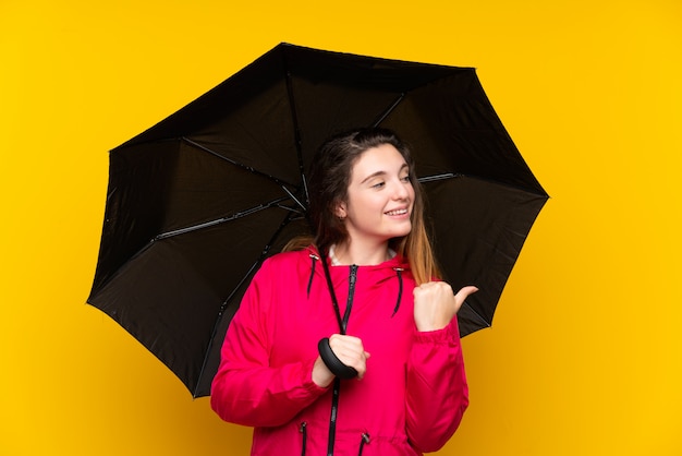 Jeune fille brune avec un parapluie sur un mur jaune isolé pointant sur le côté pour présenter un produit