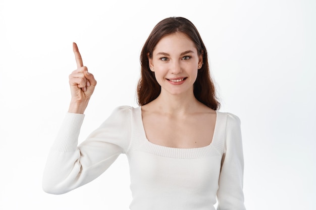 Jeune fille brune positive montrant le chemin, pointant le doigt de côté, montrant une publicité sur le dessus et souriant, faisant la promotion de la bannière de votre entreprise, debout sur un mur blanc