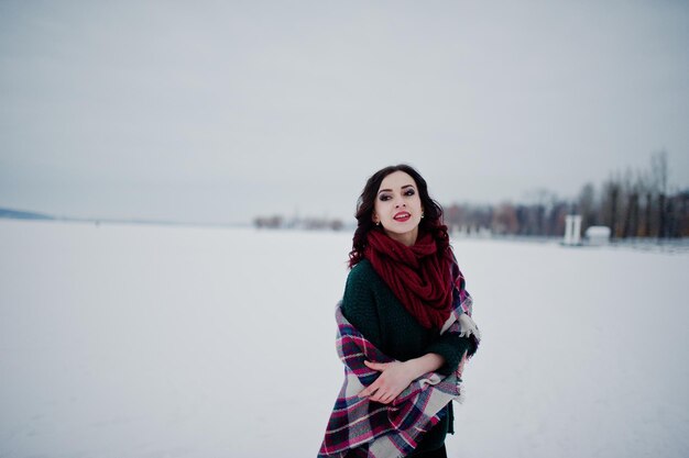 Jeune fille brune en pull vert et écharpe rouge avec lac gelé extérieur à carreaux le soir de la journée d'hiver